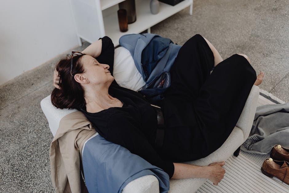 A woman rests on a sofa indoors, surrounded by casual clothing and shoes.